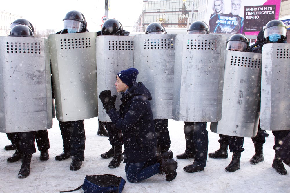 A young man kneels in the snow in front of police in combat gear behind silver shields